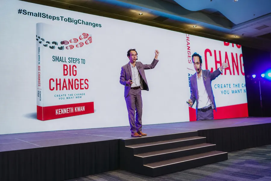 Kenneth Kwan on stage beside the Small Steps to Big Changes book cover